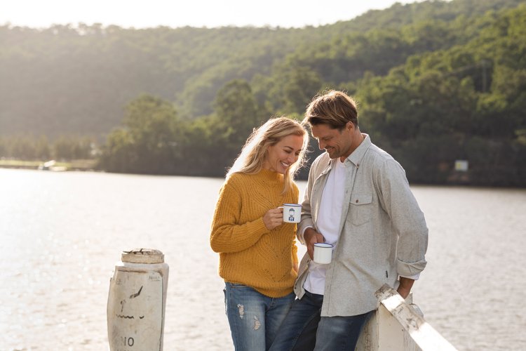 Adult couple enjoys a tranquil moment by the water, casually holding mugs while overlooking a scenic view of a green, forested hillside.