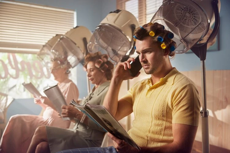 Three women in a vintage hair salon with hair dryers; one woman is talking on a cell phone, and the other two are reading magazines.