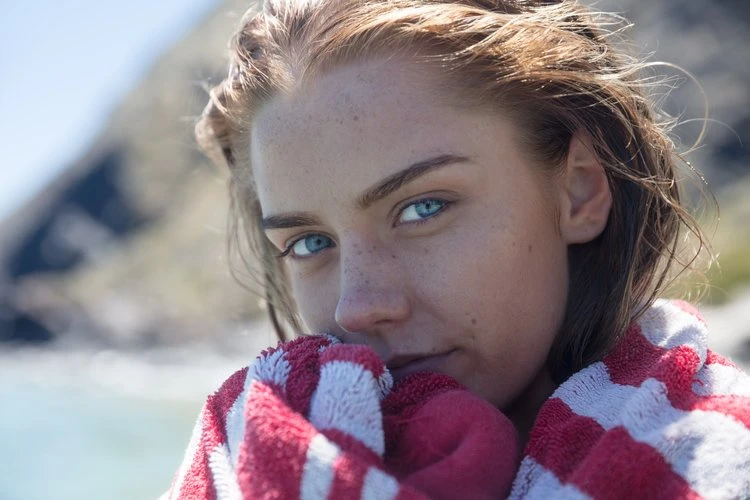 Close-up of a young woman with blue eyes, freckles, and reddish-brown hair, wrapped in a striped towel on a sunny day.