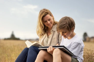 Woman and child sitting outdoors in a dry grassy field, reading a book and writing on a laptop.