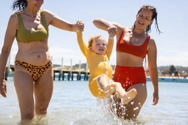 Three people in shallow coastal water including a child in a yellow rash guard being held by two women