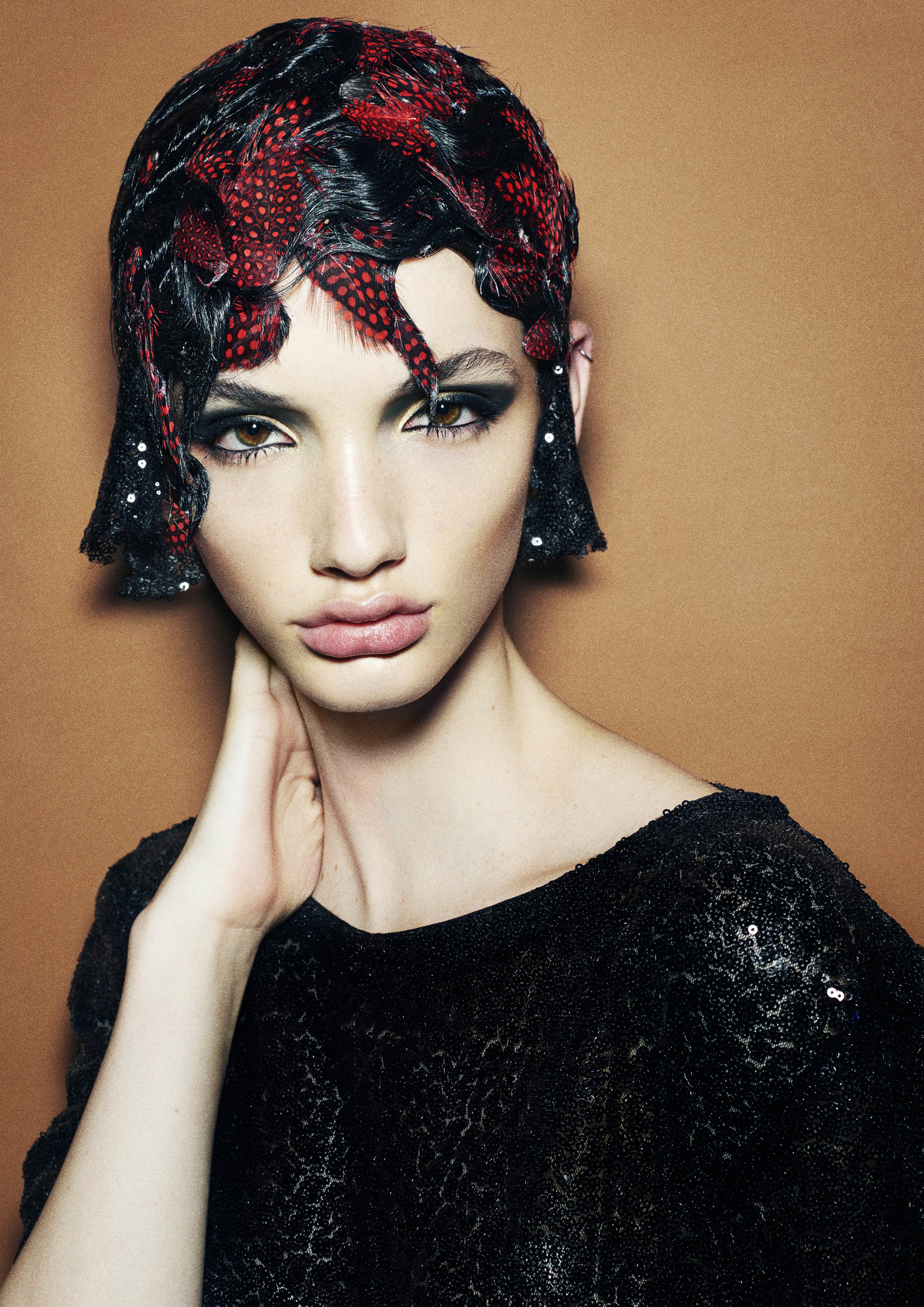 Portrait of a woman with dramatic makeup and a feather headpiece. She wears a black, sequined top against a neutral background.