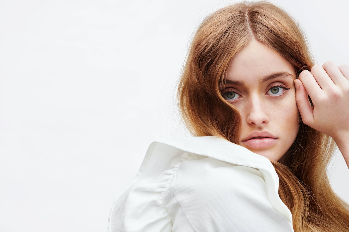 Close-up of an adult female model with long wavy red hair and light skin, wearing a white blouse, looking toward the camera. 