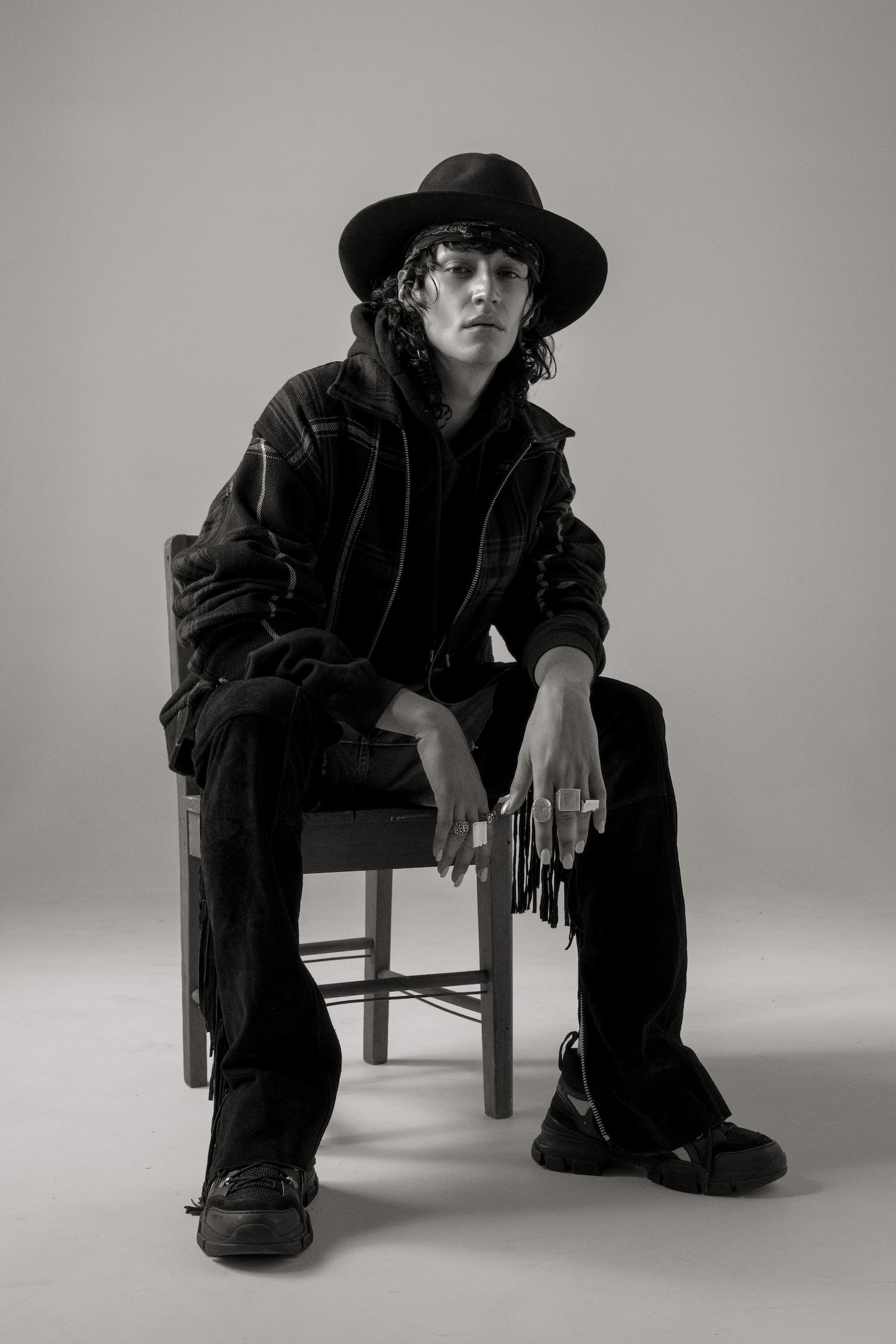 Portrait of a slender adult man wearing a hat and dark clothing, seated on a chair against a gray backdrop in a studio setting.