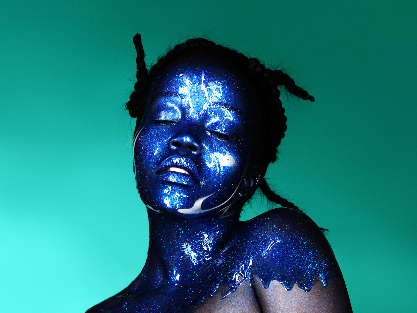 Headshot of a Black woman with glittery blue body paint and closed eyes against a teal backdrop in a studio setting. Beauty shot.