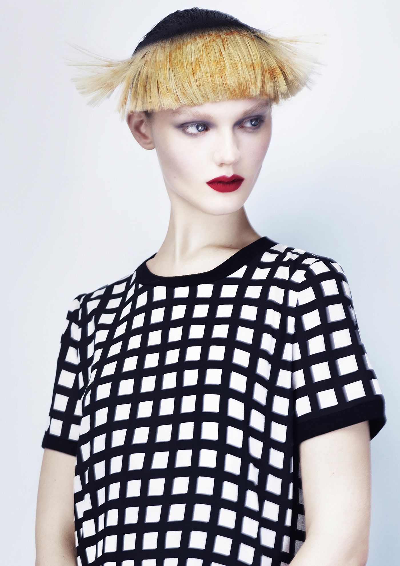 Headshot of a pale-skinned fashion model with a blonde bob hairstyle and a black and white grid-patterned shirt against a white backdrop.