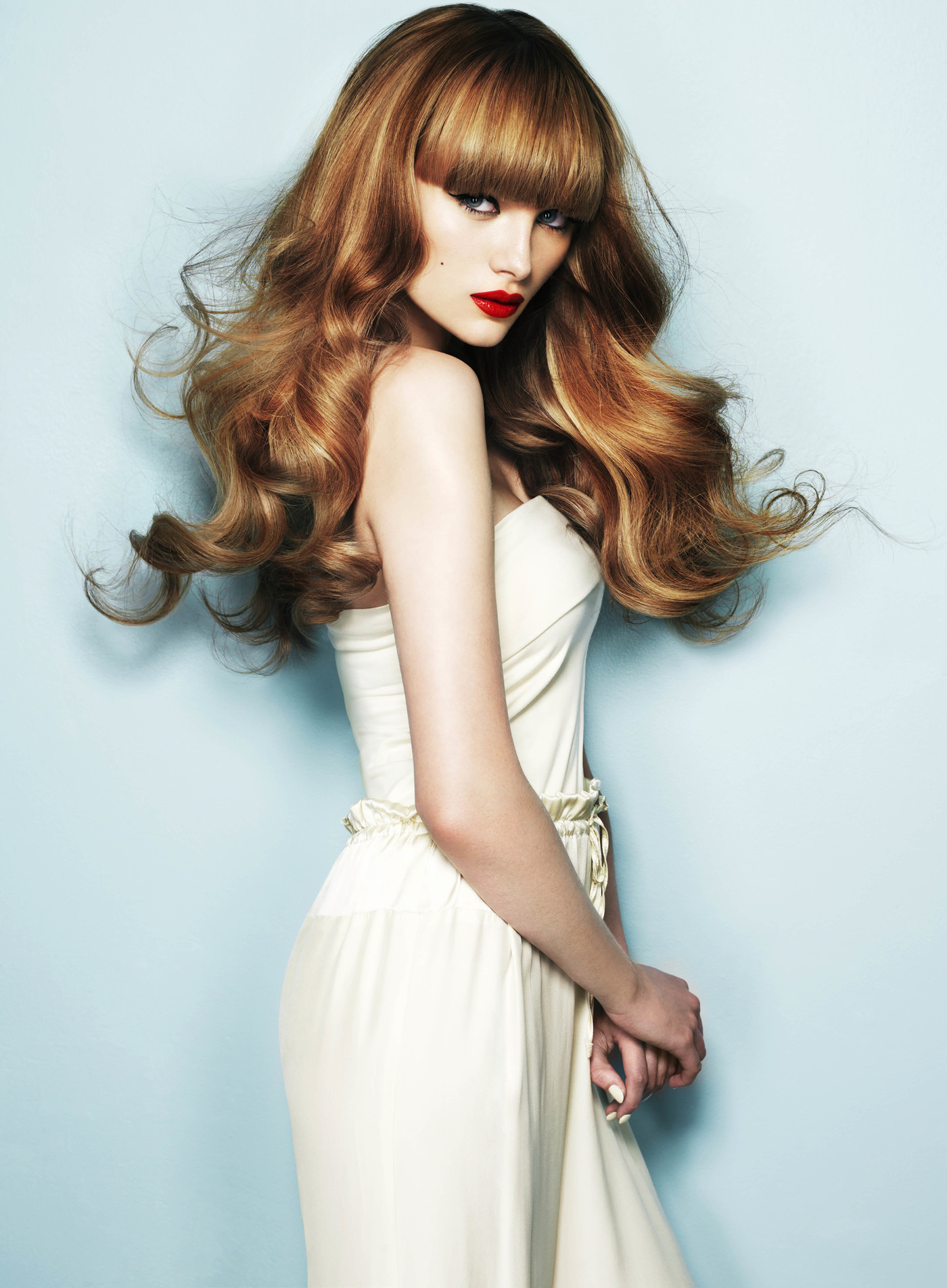 Elegant studio shot of a female model with auburn wavy hair and bangs, wearing a light dress against a soft light blue background.