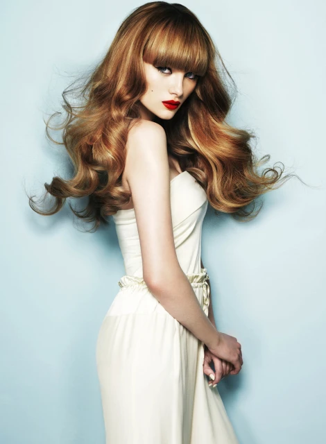 Elegant studio shot of a female model with auburn wavy hair and bangs, wearing a light dress against a soft light blue background.