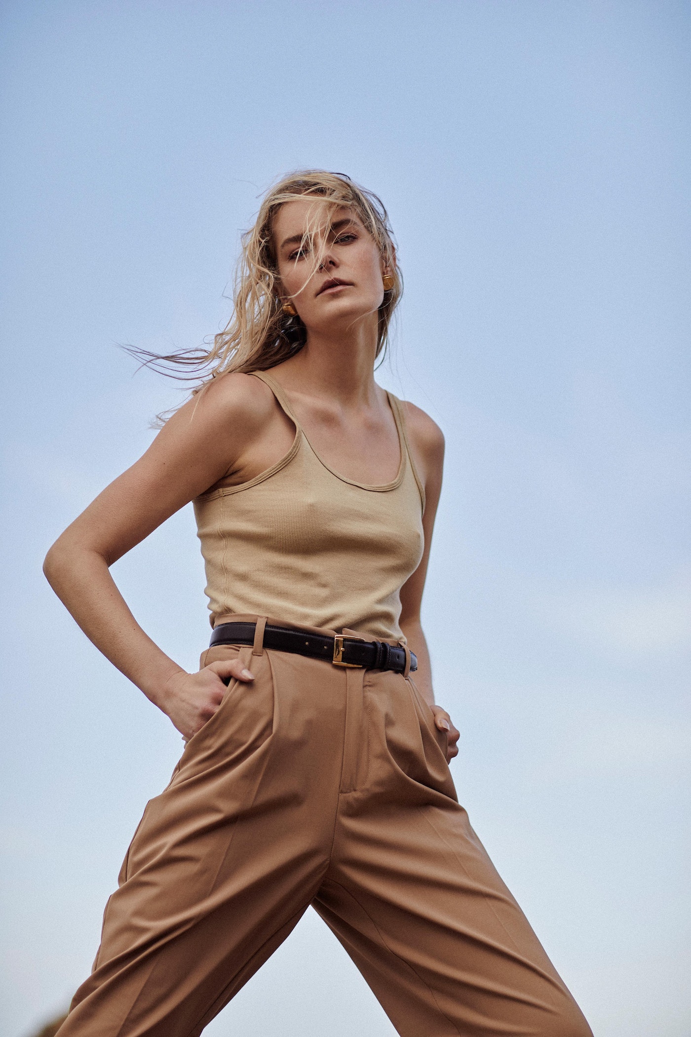 Blonde model wearing a beige tank top and brown high-waisted pants stands in a low-angle shot against a light blue sky background.