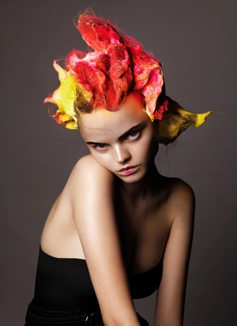 Portrait of a young woman with yellow and red styled hair, wearing a strapless black top against a neutral grey background.