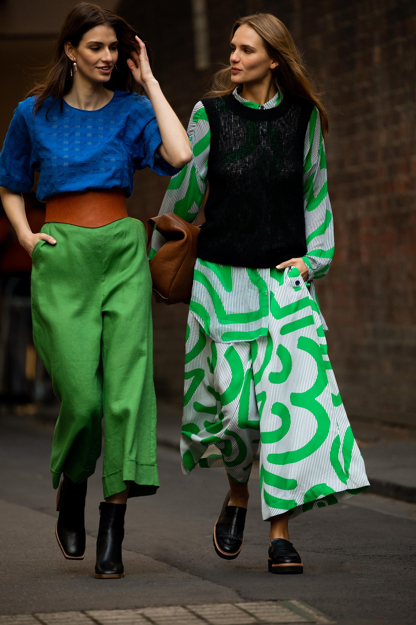 Two fashion models in bright, patterned outfits walk on city street. The model on the left wears green pants with blue top and wide belt.