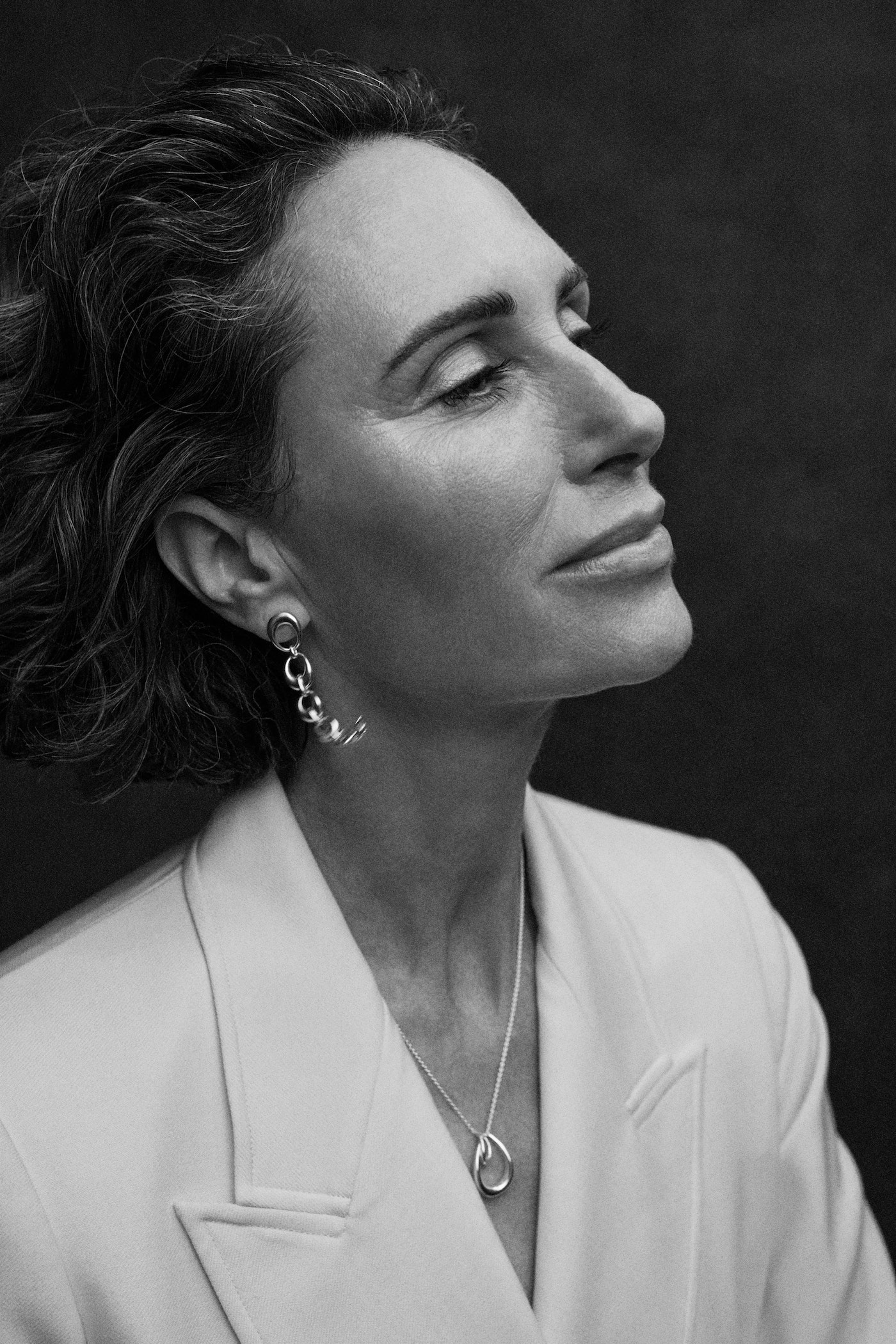 Monochrome portrait of a stylish mature woman in a cream-colored blazer, wearing statement earrings and a silver necklace, against a dark backdrop.