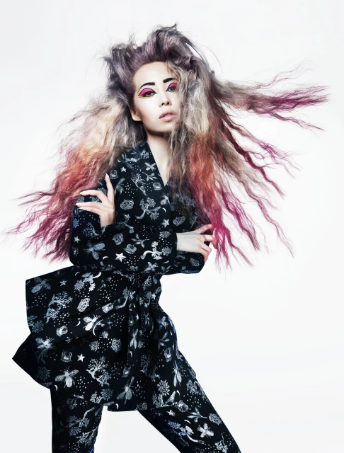 Young woman in patterned black suit with flowing, colorful dyed hair posing in a studio setting against a white background. Fashion.