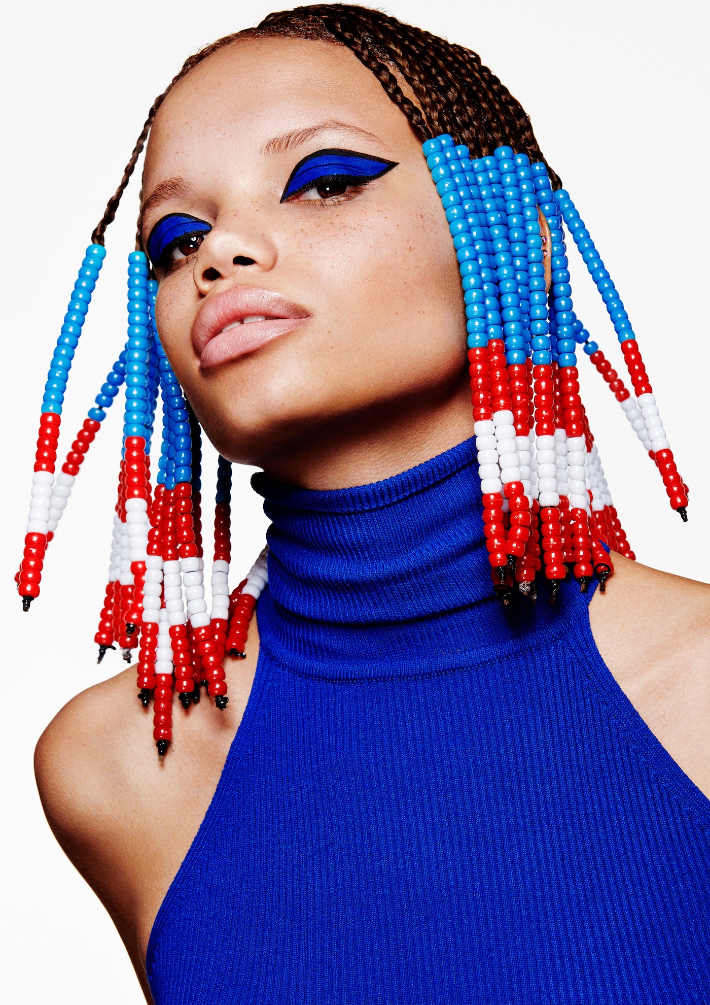 Close-up studio shot of a stylish adult female with braided hair, red, white, and blue beads, blue eye makeup, and a blue top.