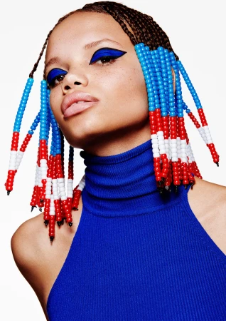 Close-up portrait of a model with braided hair decorated with blue, red, and white beads, wearing bold blue eyeliner and a blue top.