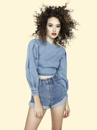 Woman with voluminous curly hair wearing a blue denim long-sleeve top and shorts against a solid cream background