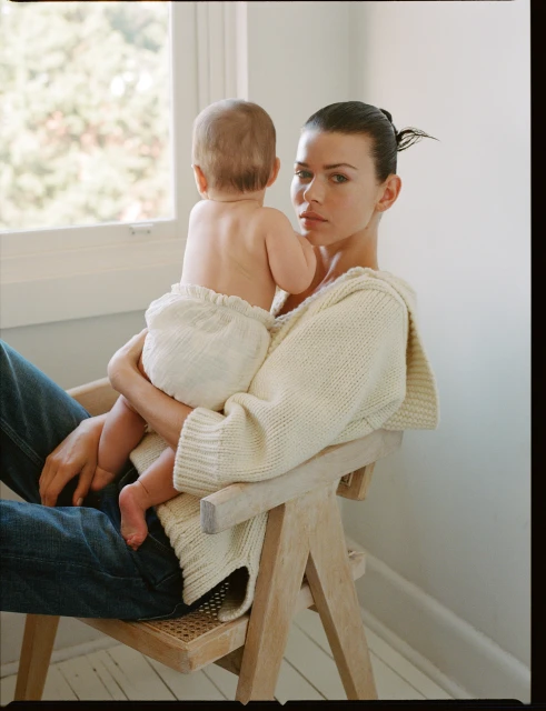 Woman holding baby in lap sitting in chair indoors near window wearing sweater and jeans. Natural light creates a soft, intimate moment.