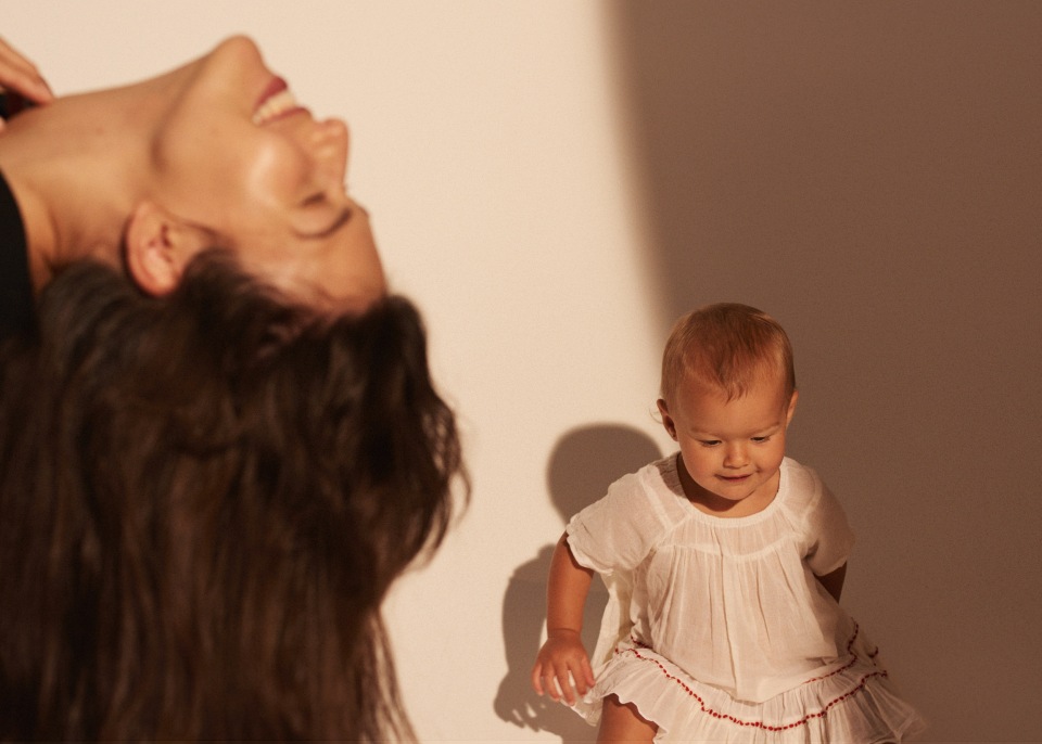 A mother with long brown hair smiles upside down near a baby wearing a white dress on a light background in a photo studio.