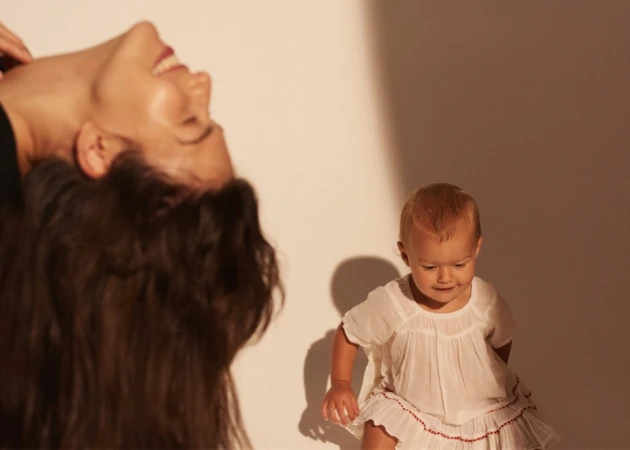 A mother with long brown hair smiles upside down near a baby wearing a white dress on a light background in a photo studio.