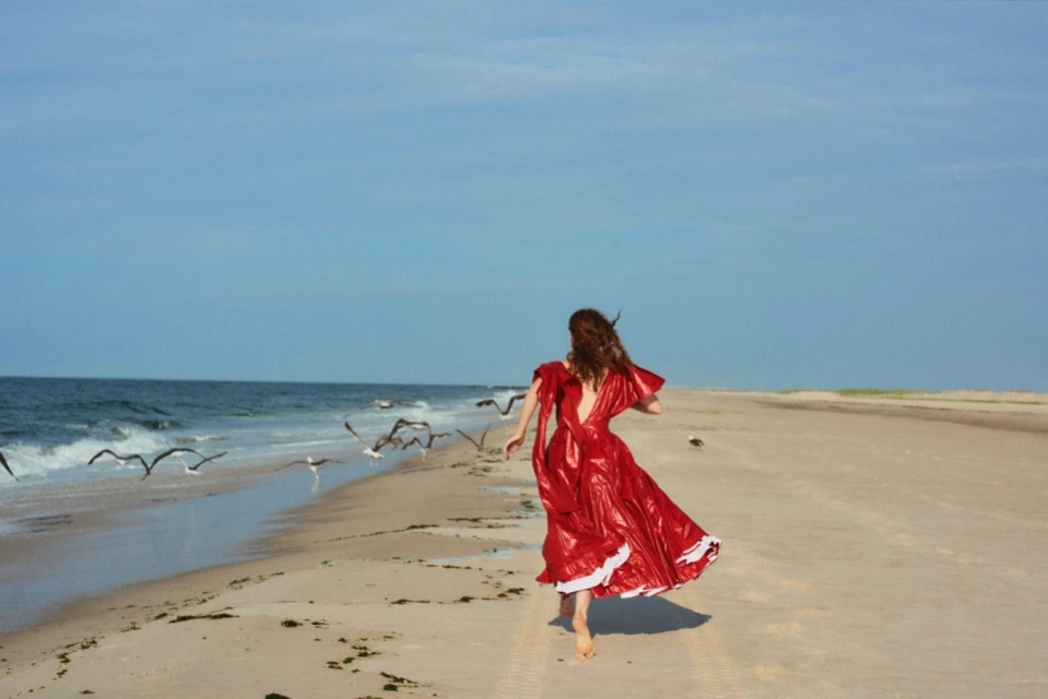 A woman in a red dress runs barefoot on a sandy beach near the ocean, with seagulls flying overhead under a clear blue sky.