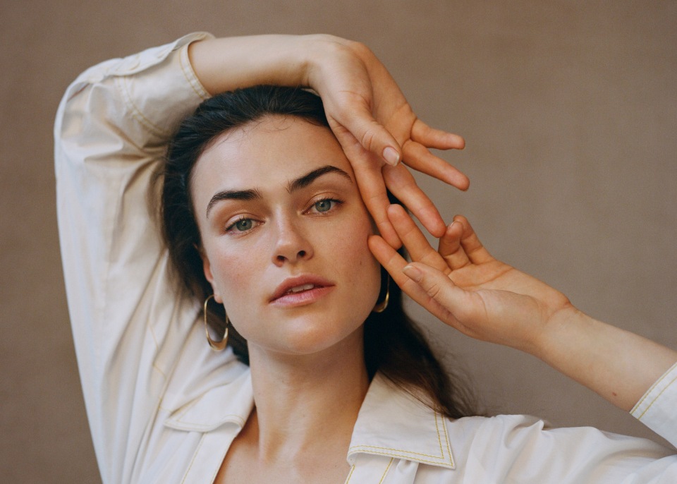 Portrait of a beautiful brunette woman with hands framing her face, wearing gold hoop earrings and a white button-down shirt against a tan backdrop.