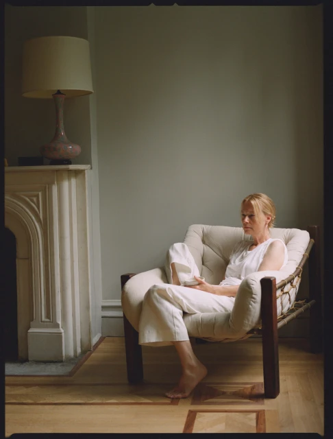 An adult woman sits barefoot in a comfortable armchair indoors, with neutral colors and natural lighting in a minimalist interior setting.
