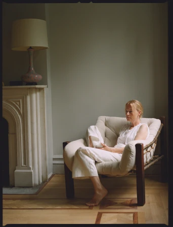 An adult woman sits barefoot in a comfortable armchair indoors, with neutral colors and natural lighting in a minimalist interior setting.