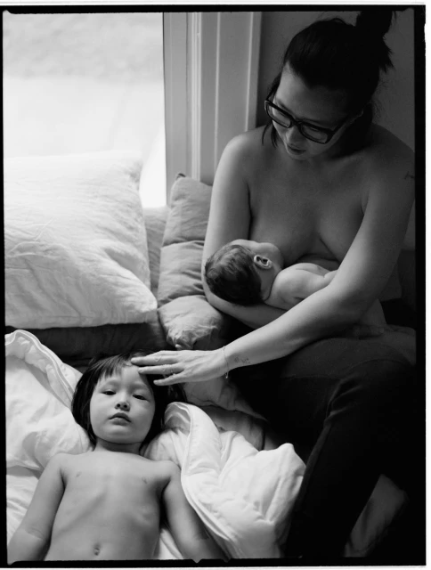 Black and white photograph of a mother breastfeeding a baby while a young child lies next to her on a bed near a window.