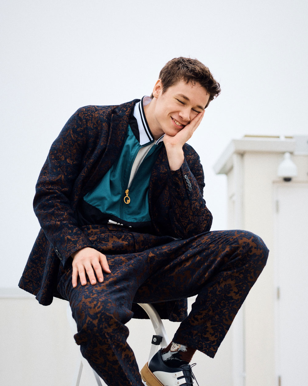 A smiling young adult male sits on a stool wearing a patterned suit with a teal jacket in an indoor setting with neutral background.