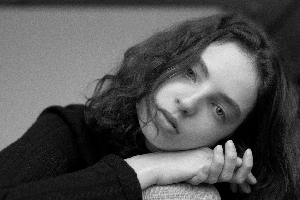 Black and white portrait of a young woman with curly hair, leaning on her hands indoors, featuring a minimalist grey background.
