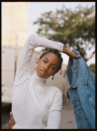 Black woman in a white turtleneck holding a denim jacket against an outdoor street background by photographer Bec Lorrimer.