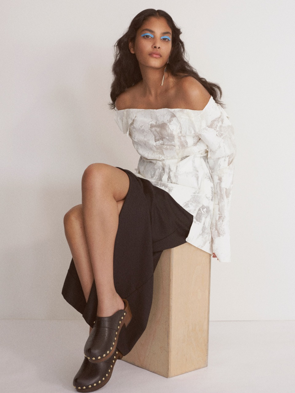 A stylish adult woman sits on a block in a photo studio wearing an off-shoulder white top, black skirt, and studded black clogs.