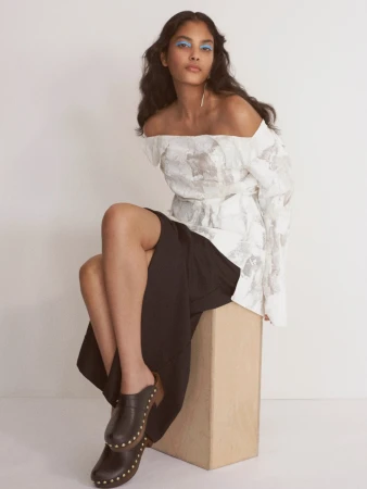 A stylish adult woman sits on a block in a photo studio wearing an off-shoulder white top, black skirt, and studded black clogs.