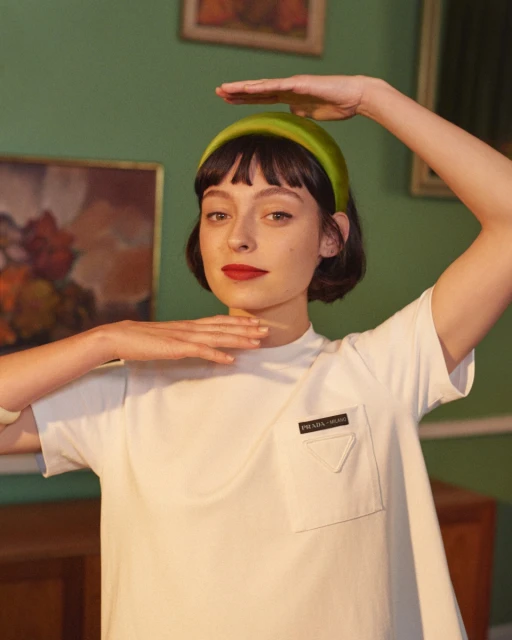 Adult female poses indoors wearing a white top and green headband with her hands raised, presenting a portrait-style composition.
