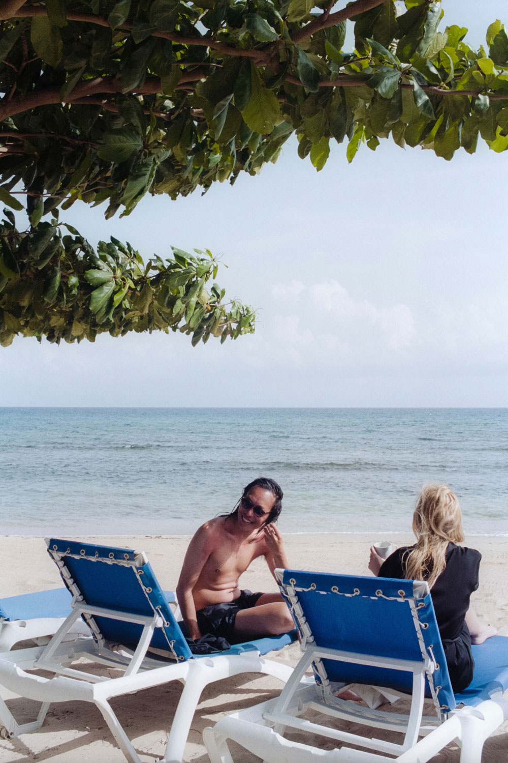 Two people relaxing on blue and white sun loungers on a sandy beach under tree branches by the ocean.