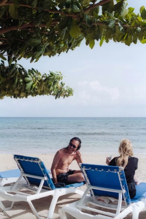 Two people relaxing on blue and white sun loungers on a sandy beach under tree branches by the ocean.