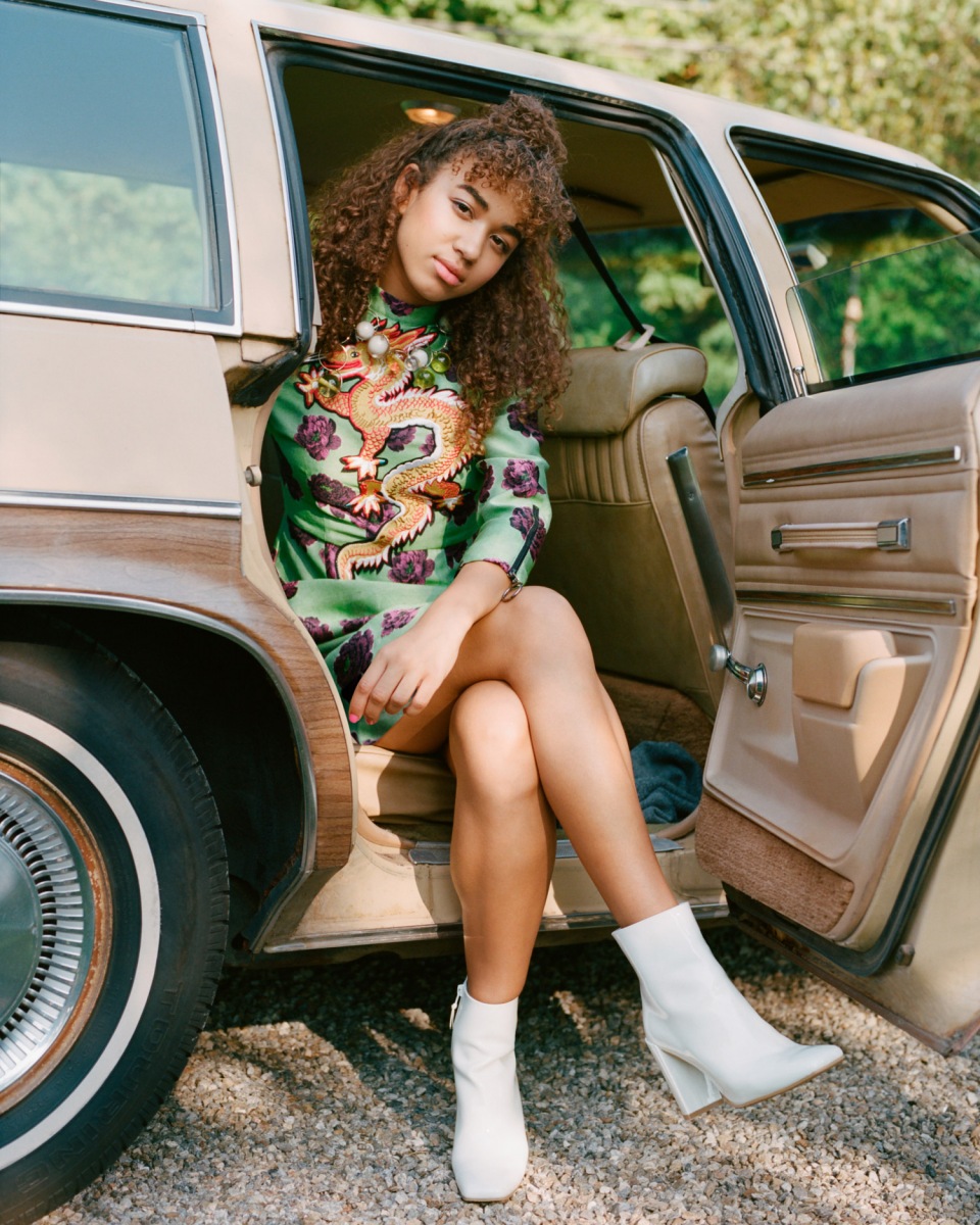 Stylish young woman with curly hair in green dress and white boots posing inside a vintage car. Natural outdoor setting.