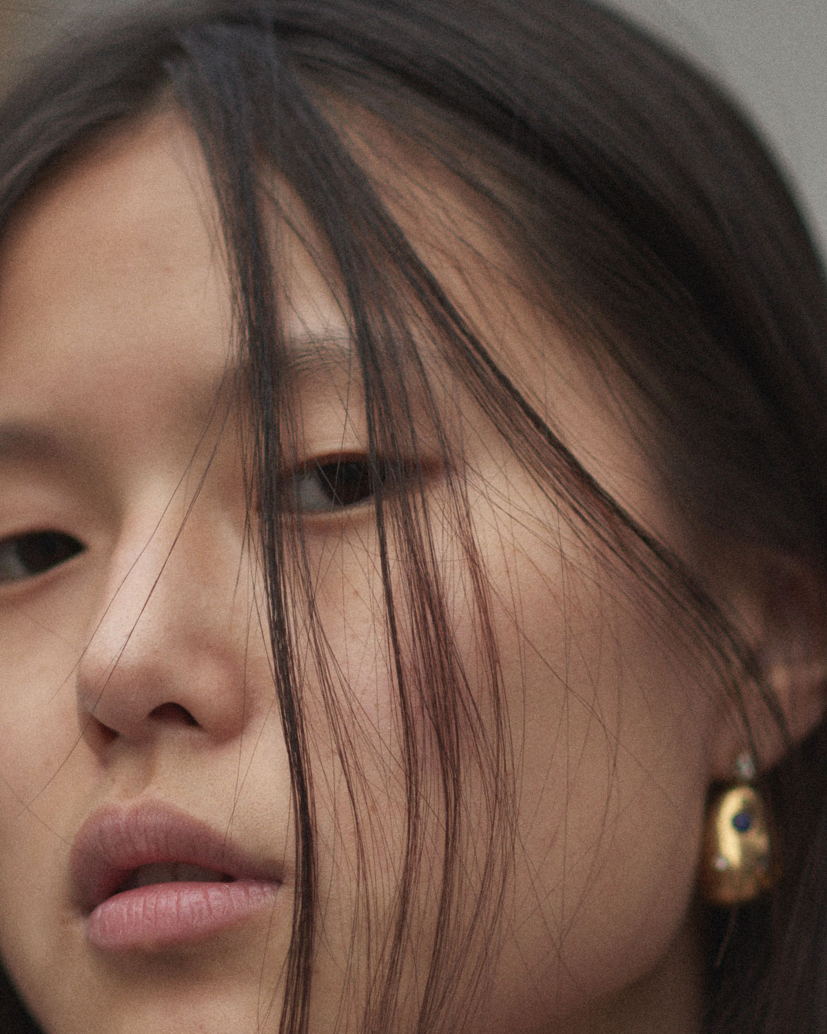 Close-up portrait of a young Asian woman with dark hair partially obscuring her face, featuring gold drop earrings with a blue stone.