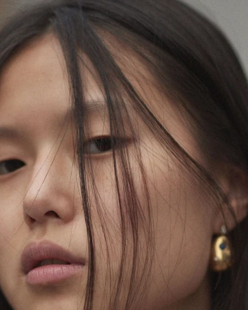 Close-up portrait of a young woman with dark hair strands across her face, wearing a gold earring with blue gemstone detail.