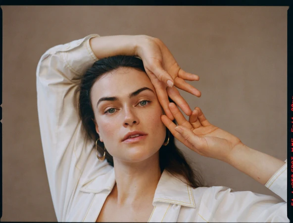 Portrait of a woman wearing a white collared shirt with her hands raised near her face against a neutral taupe background.
