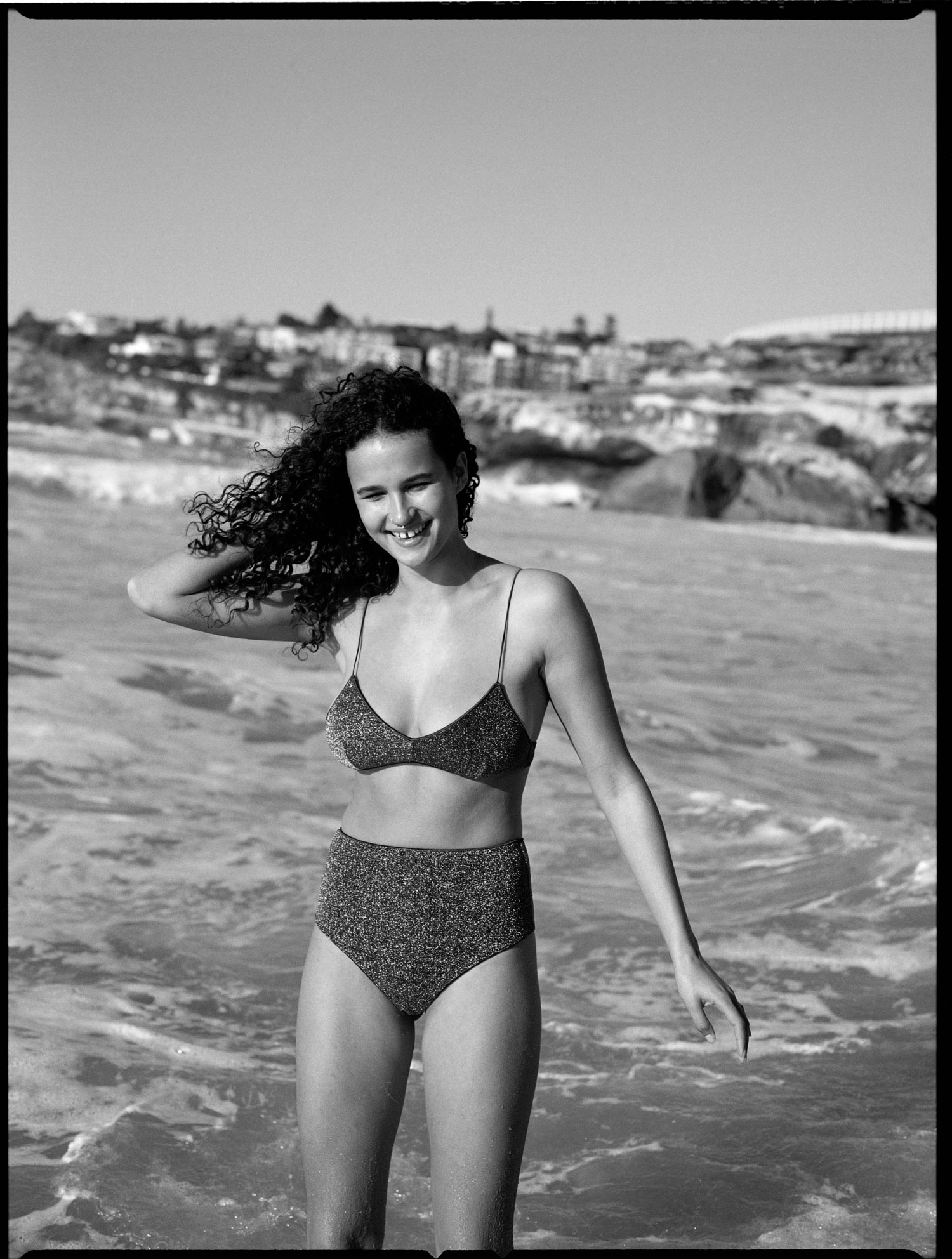 Young woman in a bikini smiles on a sunny beach with town and cliffs in the background; black and white, swimwear, curly hair.