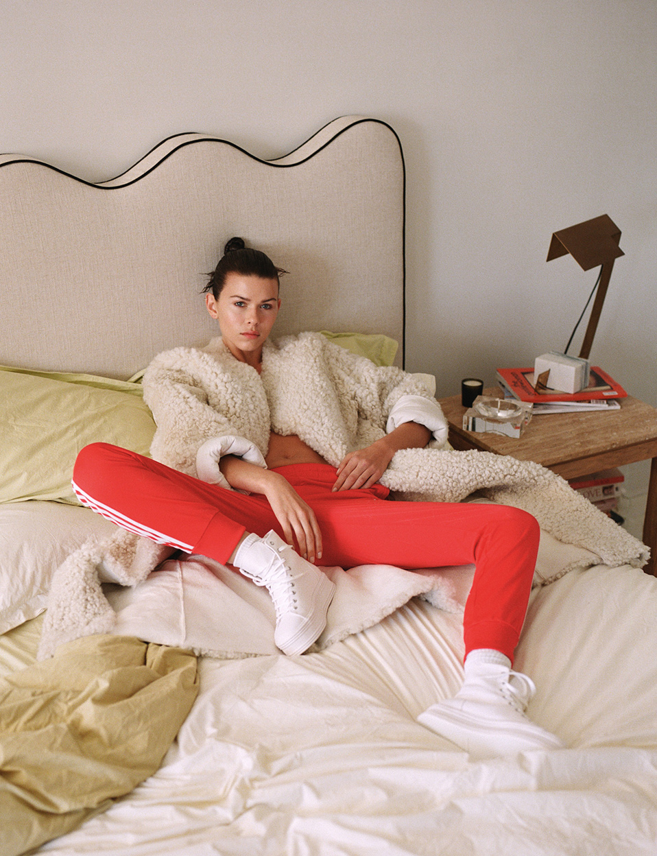 Young woman in casual outfit relaxes on bed in relaxed pose, wearing fleece jacket and red pants in a bright, inviting bedroom.