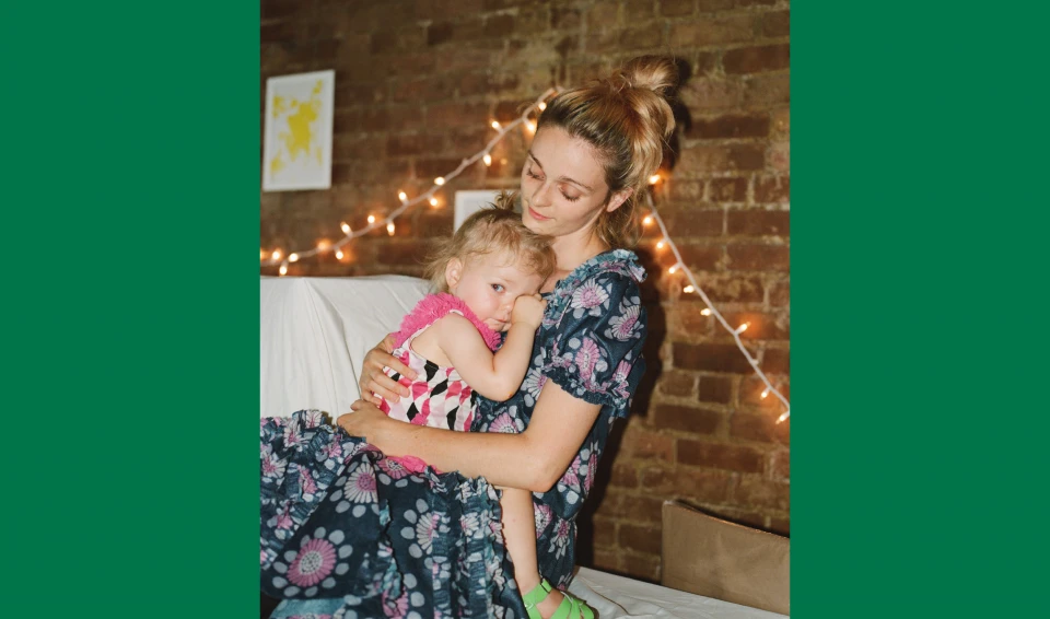 A mother holds her young child indoors near a brick wall decorated with string lights, creating a warm and intimate family portrait.