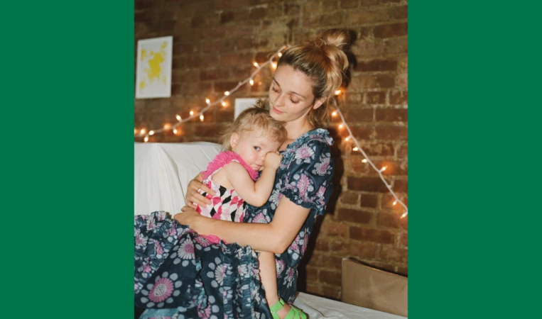 A mother holds her young child indoors near a brick wall decorated with string lights, creating a warm and intimate family portrait.