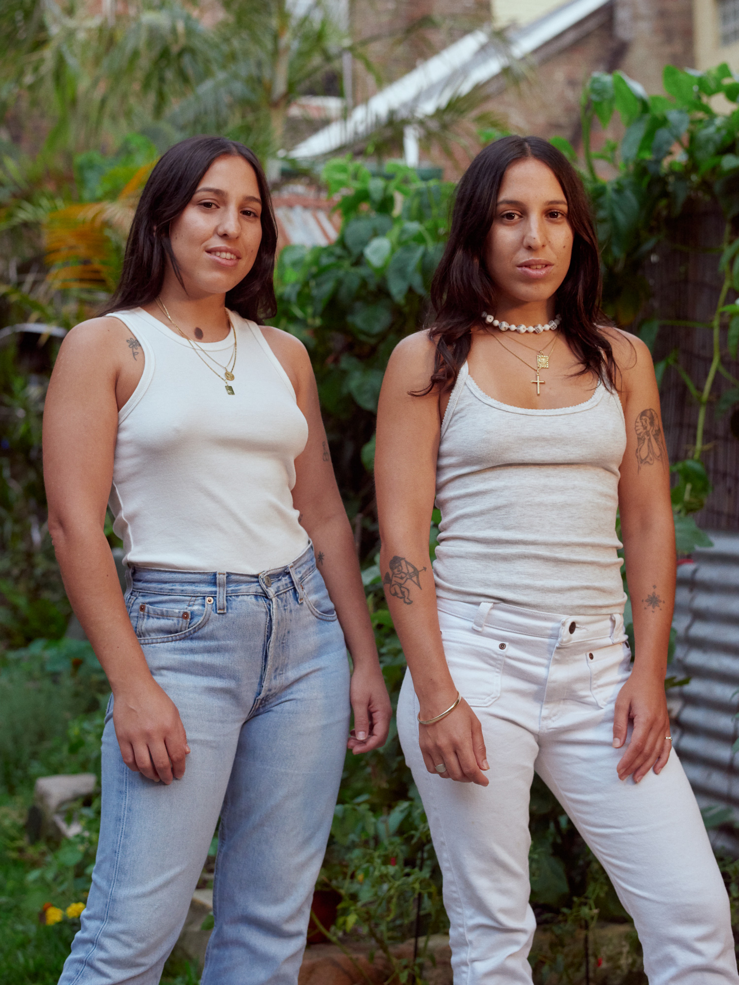 Two young women pose outdoors wearing tank tops and jeans. Lush greenery fills the background in soft, natural light.