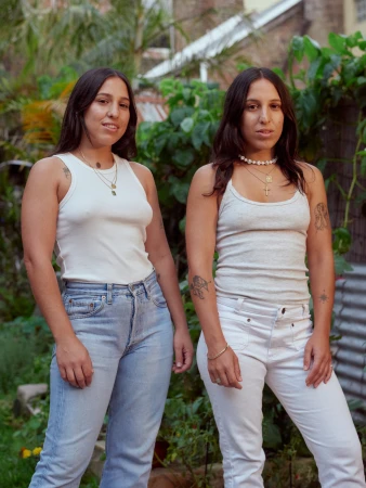 Two young women pose outdoors wearing tank tops and jeans. Lush greenery fills the background in soft, natural light.