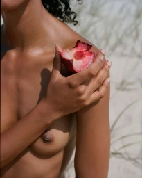 Close-up portrait of a person holding a halved red plum against their shoulder on a sandy beach by photographer Bec Lorrimer.