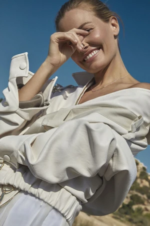 Smiling woman wearing a white leather jacket holding her hand near her face against a clear blue sky