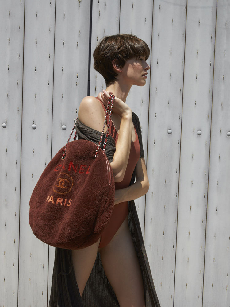 Woman in swimsuit carries a brown Chanel Paris bag in a side view against a white metal fence. Fashion shot in natural light.