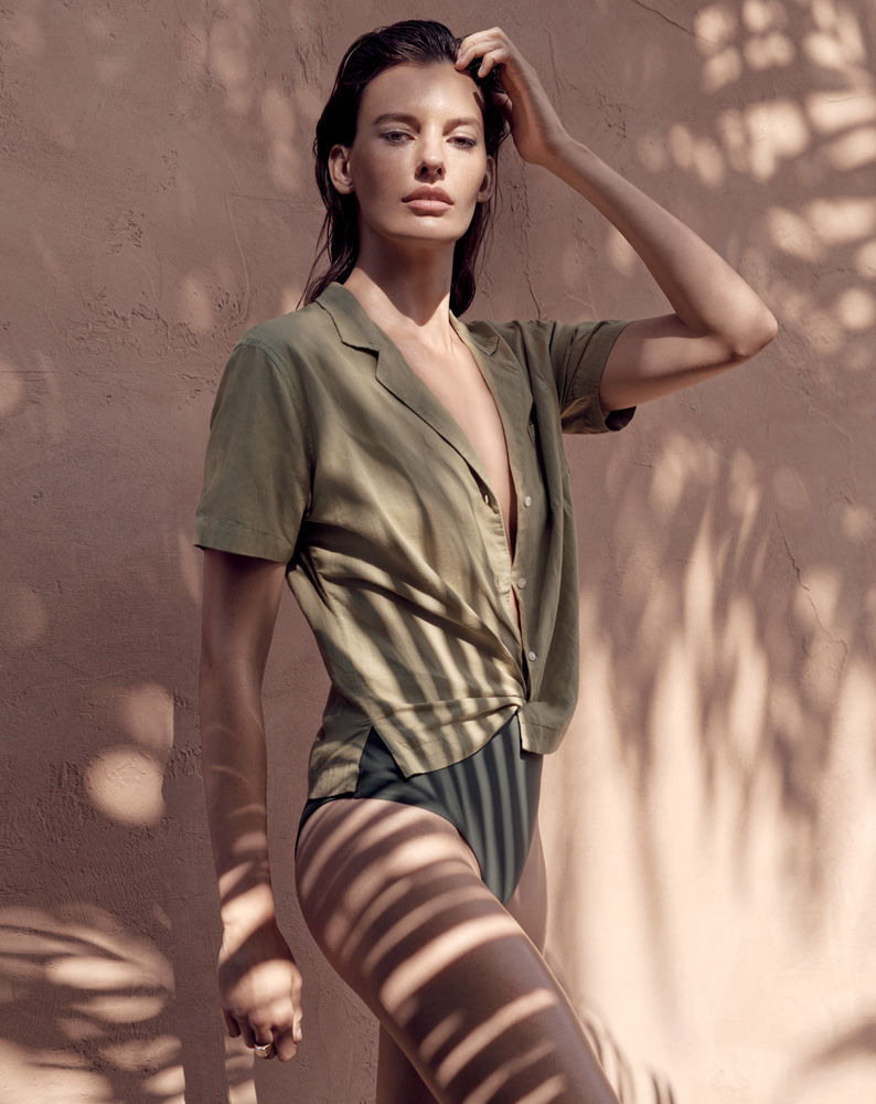 Woman poses in green shirt and black bottoms against pink wall with tropical plant shadows, fashion editorial style, sunny outdoor lighting.